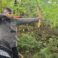 A man with gray hair is aiming a wooden bow and arrow in a forested area. The surroundings are lush and green.