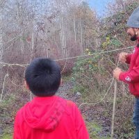 A man and a child, both in red hoodies, are outdoors setting up a rope line in a wooded area with bare trees and some bushes.