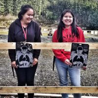 Two young women are standing outdoors in front of a wooden railing, each proudly showing their completed shooting targets.