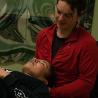 A woman in a red jacket attends to a person lying on a table, gently holding their head. The setting appears calm and professional, with indigenous art on the wall behind them.