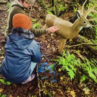 Two people in a forest setting interact with a deer-shaped target. One person kneels wearing a blue coat and orange beanie, while the other stands in a green jacket and cap, pointing at the target. Ferns and fallen leaves cover the ground around them.