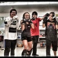 A group of six people posing in a room, dressed in boxing and martial arts attire, wearing gloves. They are smiling and appear enthusiastic.
