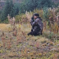 A young man is kneeling in a field, wearing a camouflage jacket and holding a long gun, surrounded by tall grass and trees in a wooded area.