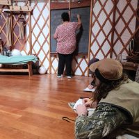 A person is writing on a chalkboard inside a yurt. Several adult students are lying on the floor, taking notes on paper.