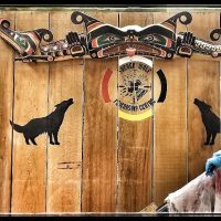 A smiling woman in an apron and blue gloves is standing in front of wooden indigenous artwork and a sign saying " Sacred Wolf Friendship Centre". She is preparing hides.